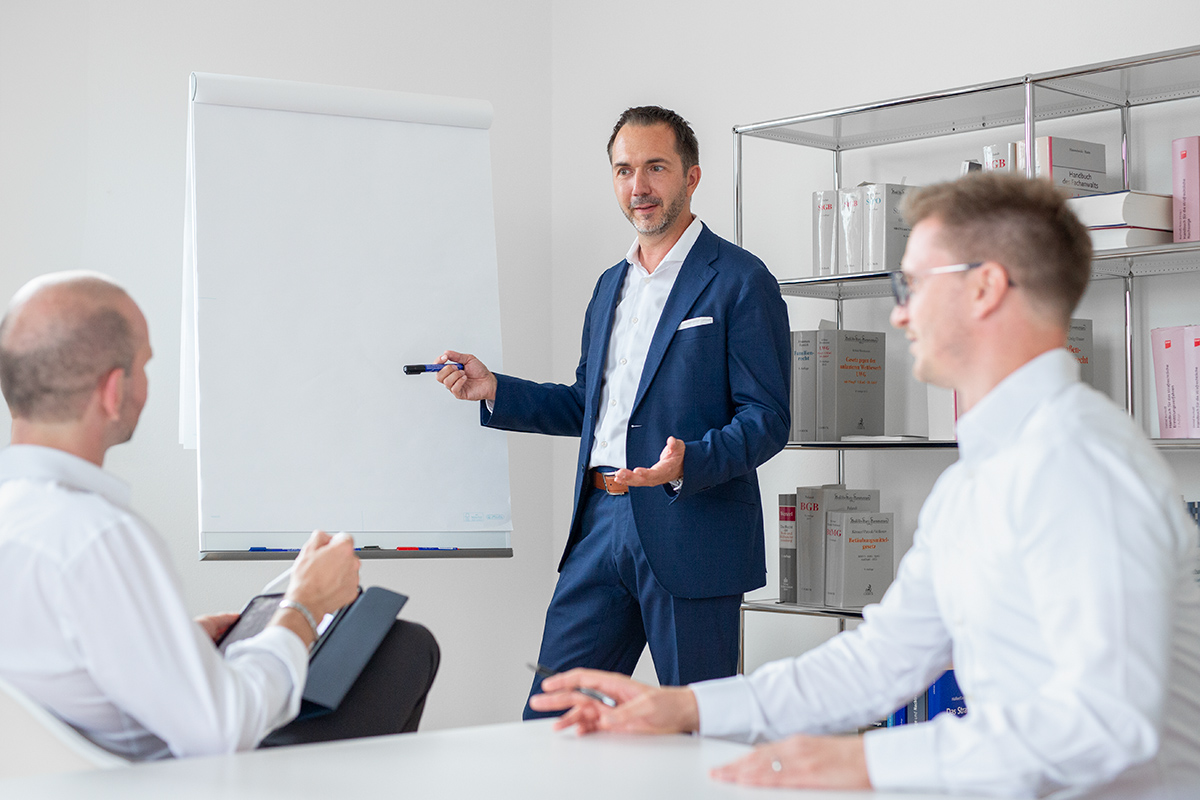 Ein Mann im blauen Anzug hält eine Präsentation an einem Flipchart vor zwei Kollegen in heller Büroumgebung