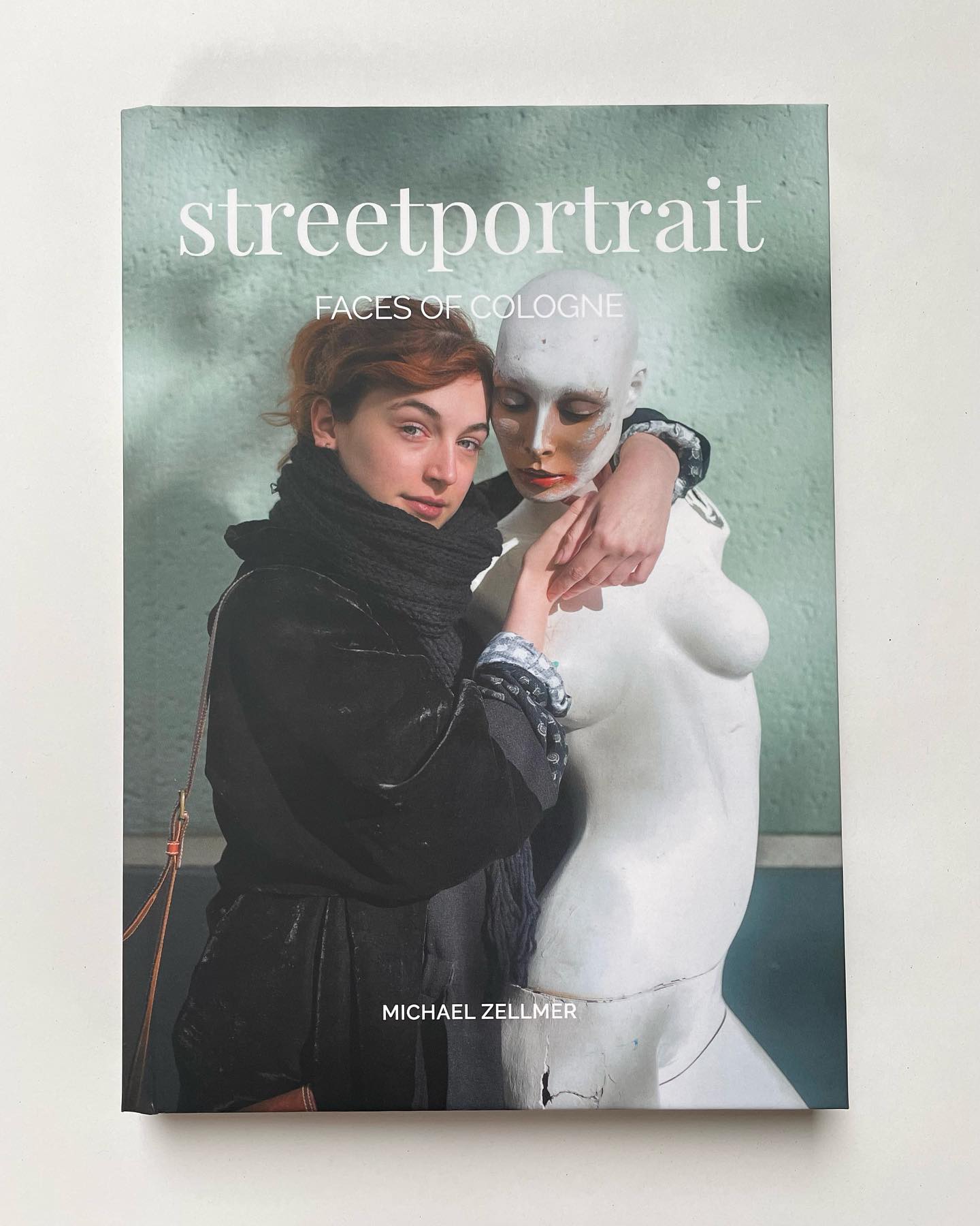 Das Buchcover "streetportrait faces of cologne" von Michael Zellmer, das eine junge Frau mit rotem Haar, Handtasche und schwarzem Schal zeigt, die eine weiße Schaufensterpuppe umarmt. Der Hintergrund ist eine grüne Wand.