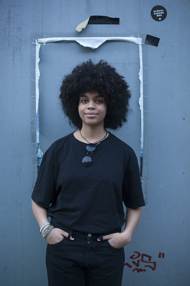 Eine junge Frau mit voller, dunkler Afro-Frisur steht in einem schwarzen T-Shirt und schwarzer Jeans. Sie hat die Hände in den Taschen und trägt eine Sonnenbrille um den Hals. Ein leichtes Lächeln liegt auf ihrem Gesicht, während sie vor einer blaugrauen Wand mit Spuren von Plakaten und Graffiti posiert.