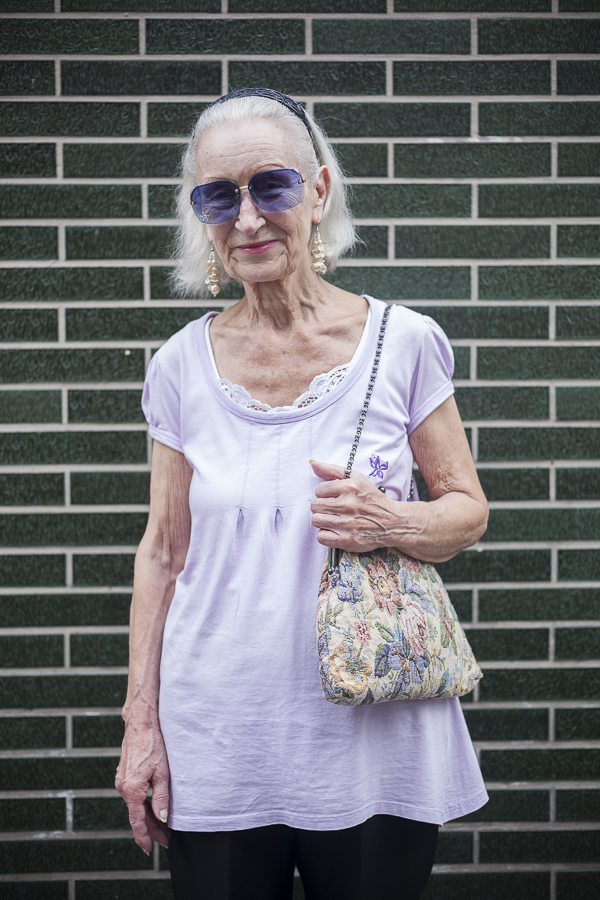 Eine ältere Frau mit weißem Haar, einer blauen Sonnenbrille, großen Ohrringen und einer bestickten Handtasche steht lächelnd vor einer dunkelgrünen Ziegelwand. Sie trägt ein hellviolettes Oberteil mit Spitze und eine dunkle Hose.