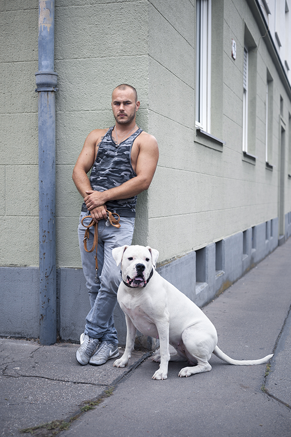Ein muskulöser Mann mit kurzen Haaren und Bart in einem Tarnmuster-Tanktop und Jeans lehnt an einer hellen Hauswand und hält eine Hundeleine in der Hand. Vor ihm sitzt ein großer, weißer Hund mit schwarzem Halsband.