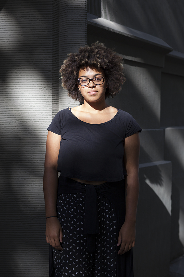 Eine junge Frau mit Afro-Frisur und Brille steht ernst und nachdenklich vor einer strukturierten grauen Wand, die teilweise von einem starken Schattenwurf bedeckt ist. Sie trägt ein schwarzes T-Shirt und ein gemustertes Kleid.