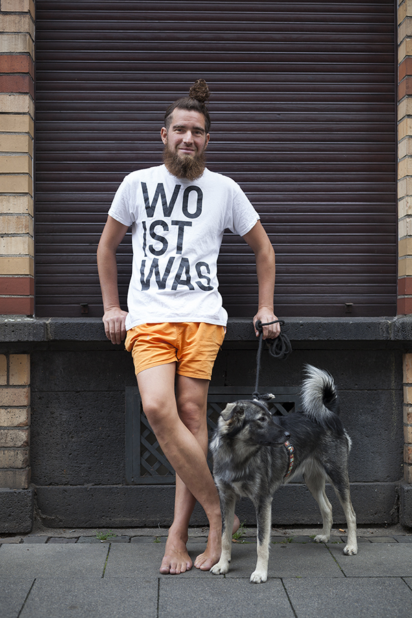 Ein junger Mann mit langem Bart, einem Zopf auf dem Kopf und orangefarbenen Shorts steht barfuß an einer braunen Rolladenwand. Er trägt ein weißes T-Shirt mit dem Aufdruck "WO IST WAS" und hält die Leine eines grauen Hundes, der neben ihm sitzt.