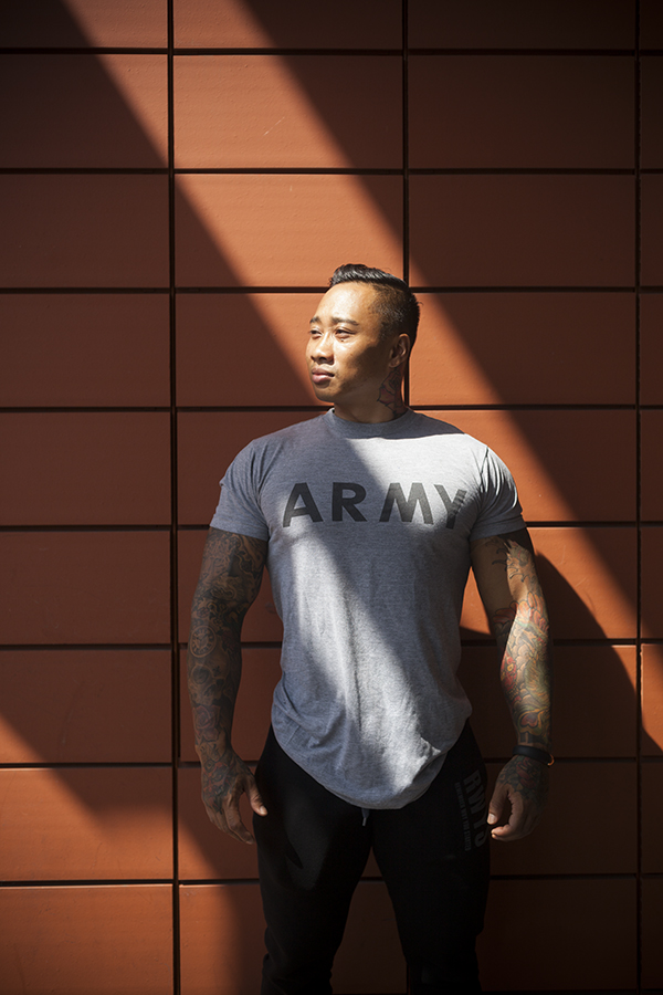 Ein muskulöser Mann mit kurzen, nach hinten gegelten Haaren und zahlreichen Tattoos an beiden Armen und am Hals steht vor einer braunen Ziegelwand. Er trägt ein graues T-Shirt mit dem Aufdruck "ARMY" und blickt nach rechts oben in das Sonnenlicht, das Schattenstreifen auf die Wand wirft.