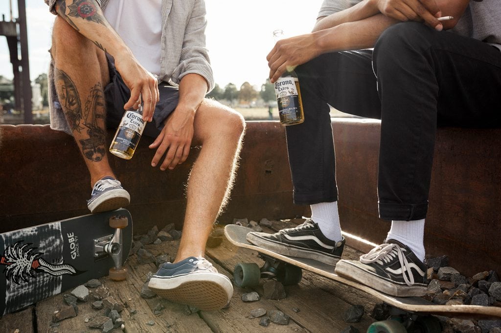 Zwei Freunde mit Skateboards und Getränken sitzen am Bahnübergang, entspannte Atmosphäre.