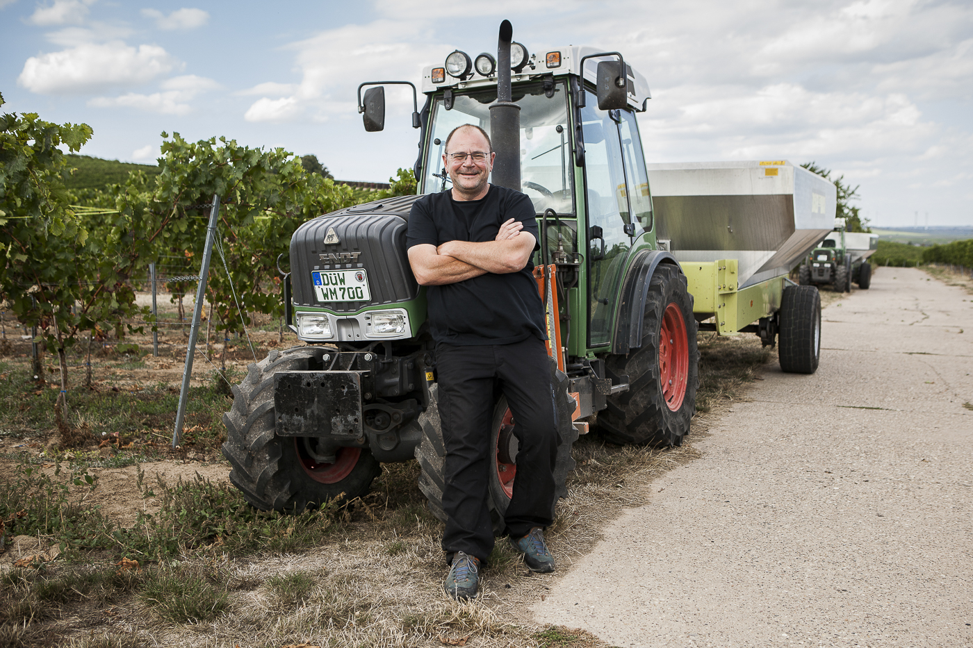 Ein Winzer mit Brille, schwarzem T-Shirt und Hose steht mit verschränkten Armen vor einem großen grünen Traktor der Marke Fendt mit der Aufschrift "Fendt WM 700". Er blickt lächelnd in die Kamera. Im Hintergrund sind Reihen von Weinreben und eine weite Landschaft unter blauem Himmel zu sehen.