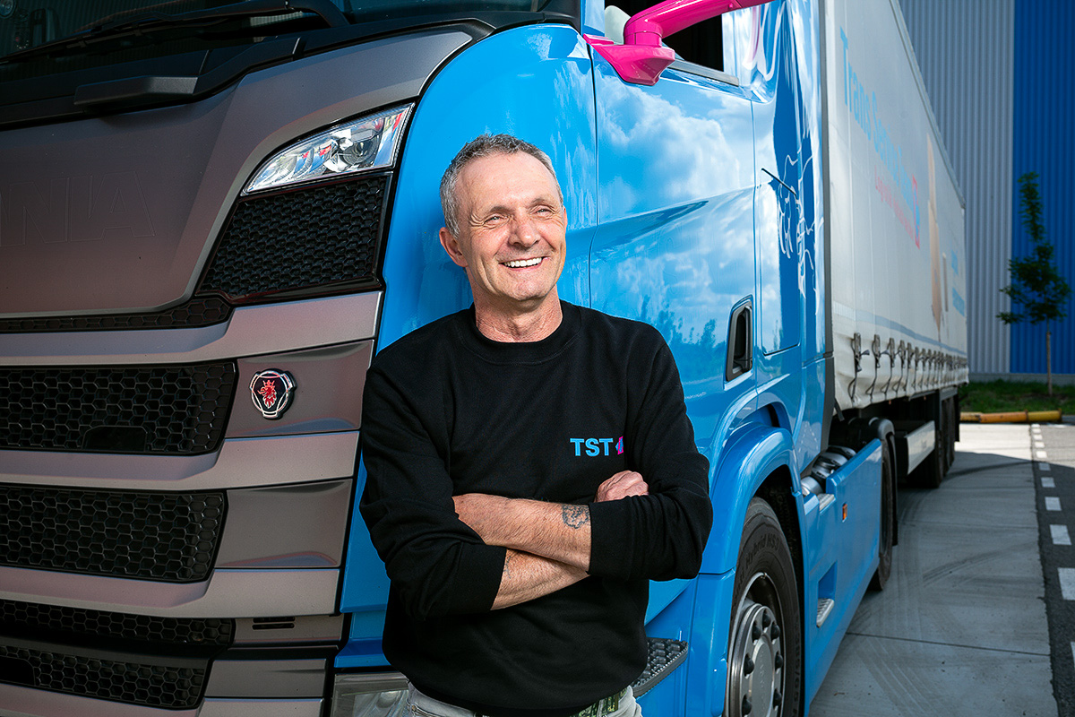 Ein Mitarbeiter eines Logistikunternehmens steht lächelnd mit verschränkten Armen vor seinem blauen LKW.