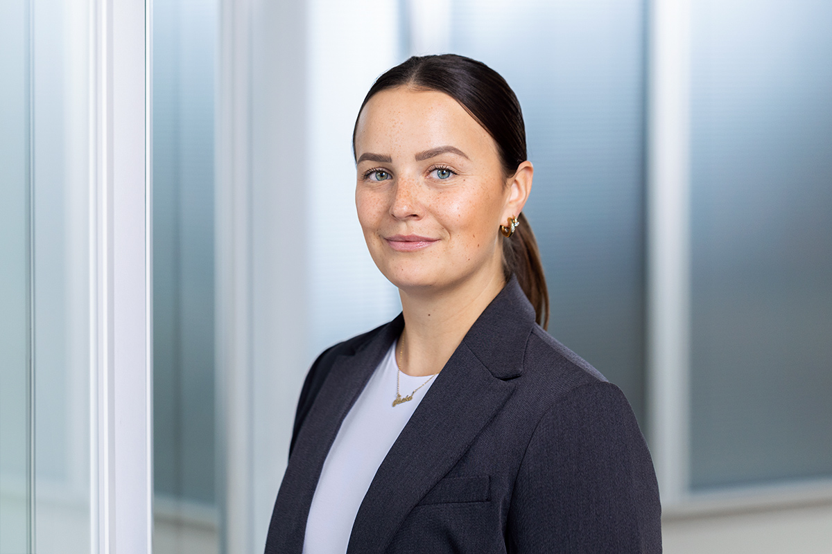 Porträt einer jungen Frau mit dunklem, zurückgebundenem Haar, Sommersprossen und blauen Augen, die ein dunkles Sakko über einem weißen T-Shirt trägt und leicht lächelnd in die Kamera blickt. Der Hintergrund ist unscharf und hell.