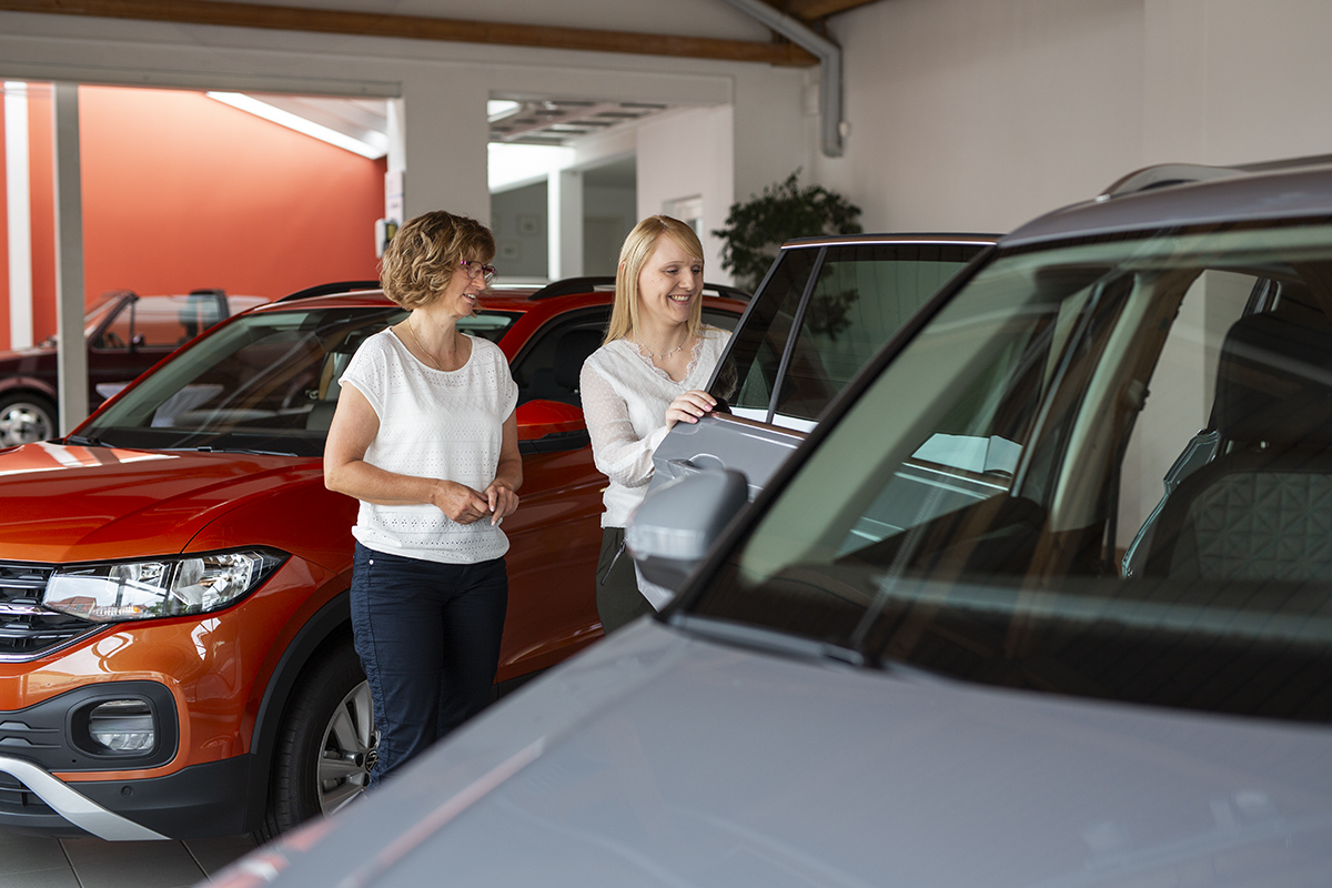 Professionelle Autoberatung: Kundin und Verkäuferin im Gespräch am Neuwagen. Im Hintergrund sind weitere helle Autos und der helle Showroom eines Autohauses zu sehen.