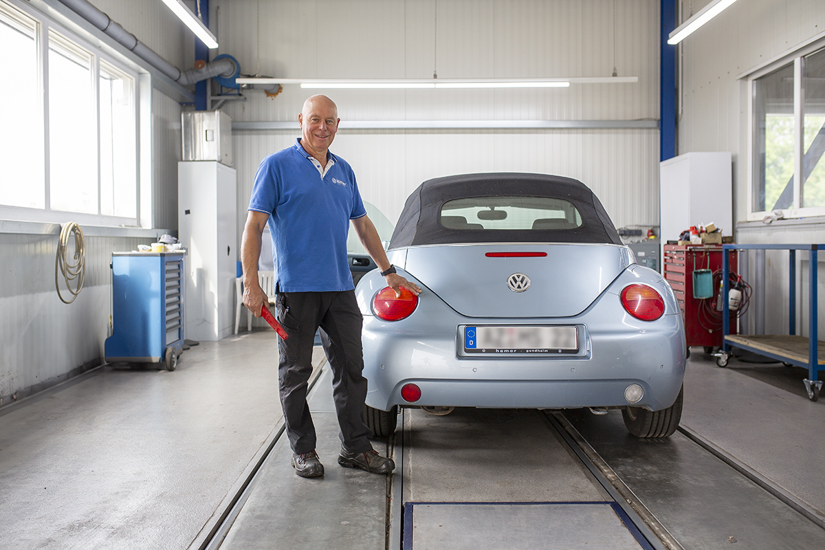 Ein lächelnder, kahlköpfiger Mann mittleren Alters in einem blauen Poloshirt und dunkler Arbeitshose steht in einer hellen Werkstatt neben einem hellblauen VW Beetle Cabriolet und hält ein rotes Werkzeug in der Hand.