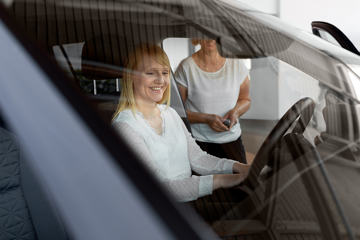 Eine lächelnde junge Frau mit blonden Haaren sitzt am Steuer eines neuen Autos und hält das Lenkrad. Hinter ihr steht eine ältere Frau mit einer hellen Bluse, die ein Objekt in den Händen hält, das einem Autoschlüssel ähnelt. Die Szene spielt in einem hellen Autohaus.