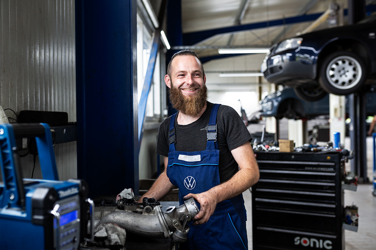 Lächelnder Kfz-Mechaniker mit Vollbart in blauer VW-Arbeitskleidung in einer professionellen Werkstattumgebung