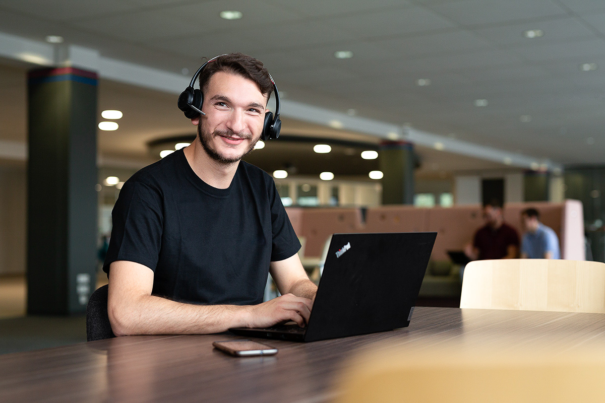 Corporate Porträt eines freundlichen jungen Mannes mit Bart und Headset, während er an einem schwarzen Laptop arbeitet, an einem Holztisch sitzend. Im Hintergrund sind unscharf weitere Personen und Büroeinrichtung zu erkennen.
