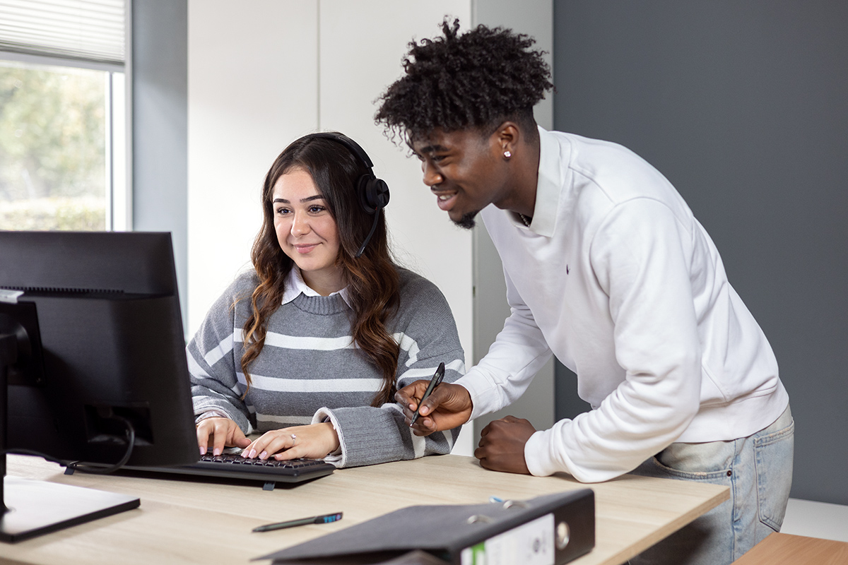 Zwei junge Auszubildende unterschiedlicher Herkunft arbeiten gemeinsam an einem Computer, symbolisieren Teamwork und Unterstützung im Büro.